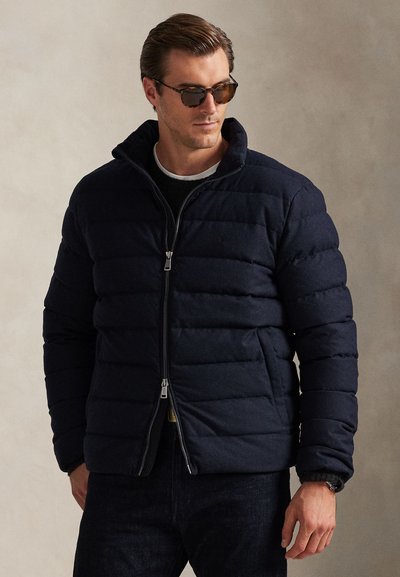 Marineblaue Steppjacke mit Reißverschluss, hohem Kragen, gestepptem Muster und Seitentaschen. Kombiniert mit dunklen Jeans und Sonnenbrille.