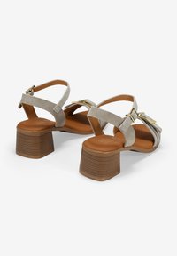 Sandalias de ante gris con tacón cuadrado, que presentan dos correas entrelazadas y acentos de hardware en tonos dorados. Plantilla lisa en marrón con suela estriada.