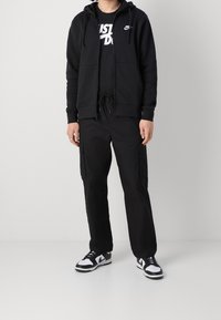 Sudadera negra con cremallera y bolsillos tipo canguro, combinada con una camiseta negra con texto blanco. Pantalones cargo negros y zapatillas en blanco y negro.