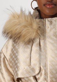 Plašč z zlatim vzorcem z umetnim krznenim obrobom kapuce, ki se ponaša z teksturirano tkanino, zategovanjem na vrvico in subtilnim sijajem.