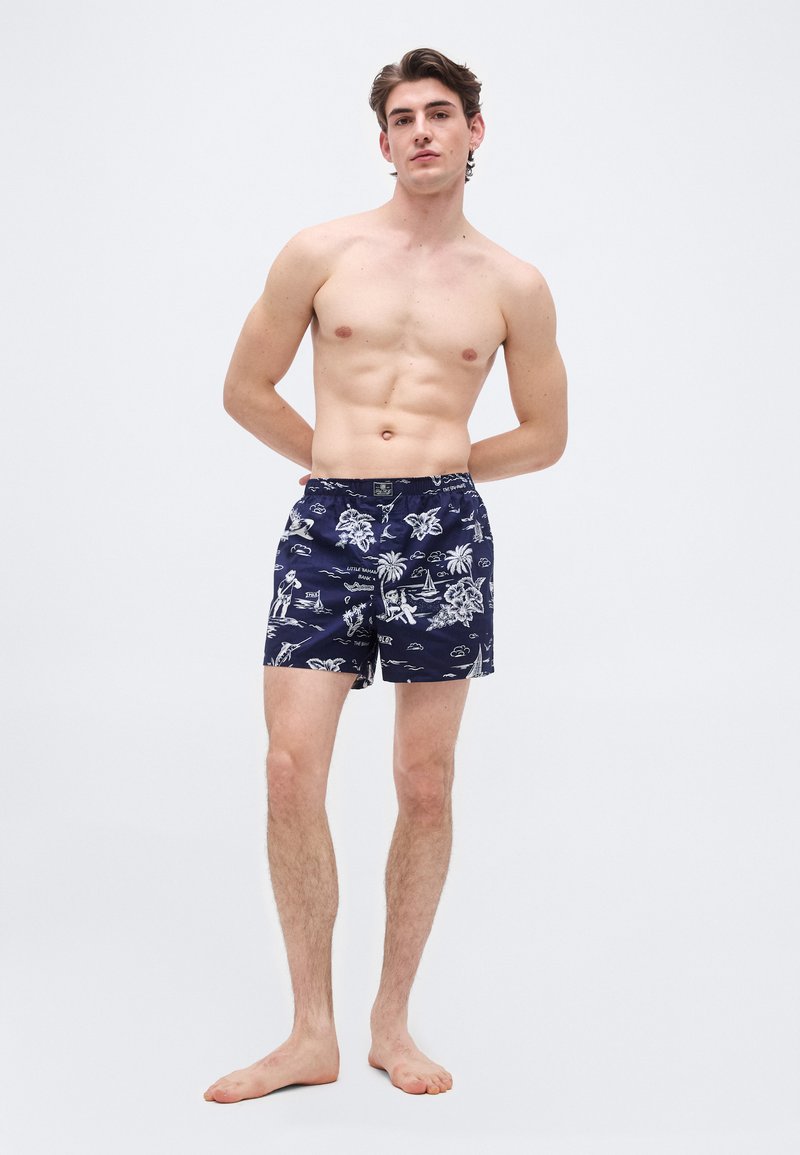 Νεαρός άνδρας που στέκεται ξυπόλητος με τα χέρια πίσω από την πλάτη, φορώντας ναυτικό μπλε μαγιό με λευκά τροπικά και ζωάκια σαφάρι.