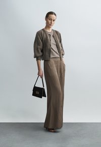 Bruine gebreide cardigan met pofmouwen, beige top en bruine wijde broek. Model houdt een zwarte handtas met metalen accent vast.