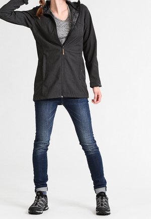 Personne portant une veste noire zippée, un t-shirt gris, un jean bleu et des chaussures de randonnée noires, debout les jambes écartées et un bras levé.