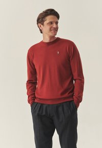 Pull rouge en maille avec manches longues, col rond et petit logo brodé sur la poitrine. Associé à un pantalon foncé plissé.