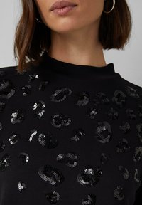 Schwarzer Sweatshirt mit einem ganzflächigen Muster aus glänzenden schwarzen Pailletten im Leopardenmuster, mit hohem Halsausschnitt und weichem Material.