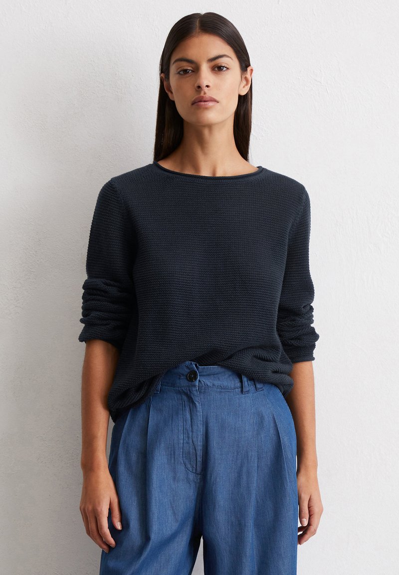 Navyfarbener Strickpullover mit strukturiertem Muster, rundem Ausschnitt und hochgekrempelten Ärmeln, kombiniert mit hellblauen, weit geschnittenen Hosen mit Knopfdetails.