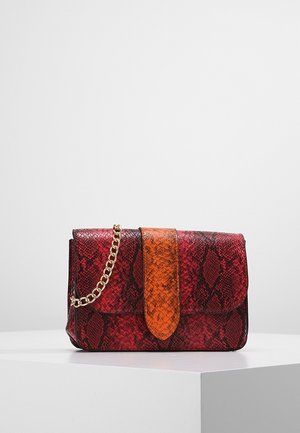 Bolso con patrón de piel de serpiente en rojo y negro, con correa naranja y cadena dorada, exhibido sobre un pedestal blanco contra un fondo gris.
