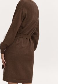 Vestito marrone lavorato a maglia con maniche lunghe, vita con cintura e polsini a coste. Presenta una vestibilità morbida e una texture soffice.