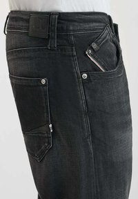 Jean en denim noir avec coupe droite, plusieurs poches et un éclat subtil. La ceinture comprend un patch en cuir griffé.