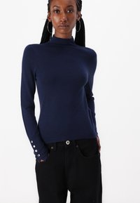 Pull noir à manches longues avec un col haut, en matière douce, avec des accents de boutons argentés aux poignets, associé à un jean noir.