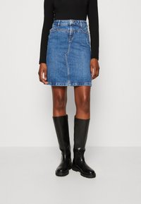 Kvinna klädd i en blå denim minikjol, svart långärmad topp och knähöga svarta läderstövlar, stående mot en oskriven vit bakgrund.