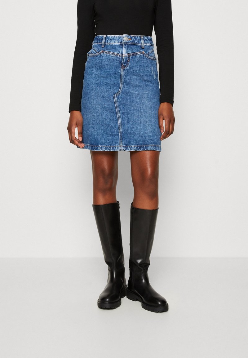 Kvinna klädd i en blå denim minikjol, svart långärmad topp och knähöga svarta läderstövlar, stående mot en oskriven vit bakgrund.