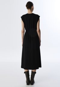 Zwarte mouwloze jurk met een geplooide taille, die tot halverwege de kuit valt. Gladde stof en minimalistisch ontwerp, gecombineerd met bruin kniehoge laarzen.