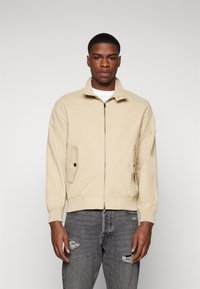 Chaqueta bomber beige con cuello alzado, cierre frontal y bolsillos laterales. Llevada sobre una camisa blanca, combinada con jeans grises desgastados.