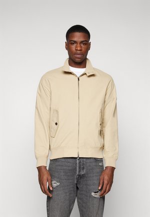 Giacca bomber beige con colletto alto, zip frontale e tasche laterali. Indossata sopra una camicia bianca, abbinata a jeans grigi strappati.