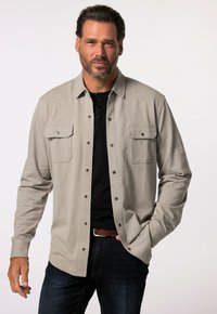 Camicia grigia chiaro con bottoni, due tasche sul petto, maniche lunghe e una texture liscia, indossata sopra una maglietta henley nera.