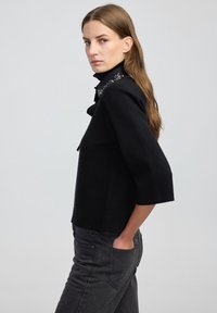 Vrouw in een zwarte top met driekwart mouwen en versierde kraag, met donkere high-waisted jeans, staande met één hand in de zak, neutrale achtergrond.