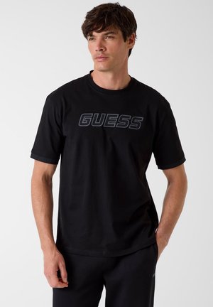 Junger Mann trägt ein schwarzes T-Shirt mit dem "GUESS"-Logo, steht mit einer Hand in der Tasche vor einem weißen Hintergrund.