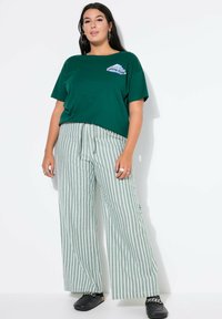 Vihreä puuvillainen t-paita, jossa on "DREAM TEAM" -grafiikka, yhdistettynä leveälahkeisiin, vaaleanvihreisiin raidallisiin housuihin ja mustiin slip-on-kenkiin.