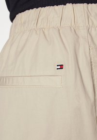 Shorts beiges avec une taille élastique, présentant une poche arrière et un petit logo brodé en rouge, blanc et bleu près de la poche.