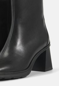 Botină din piele neagră, cu un finisaj lucios, fermoar pe lateral și toc geometric de tip bloc. Suprafață texturată, siluetă elegantă și talpă robustă.