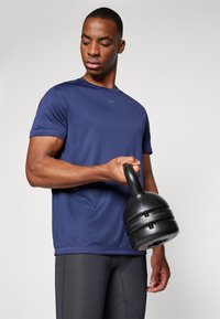 Jūras spēku īsām piedurknēm sporta krekls, izgatavots no gludas auduma, valkāts ar pieguļošām melnām legingām, turot melnu kettlebellu. Vienkāršs dizains, minimāla zīmola atpazīstamība.
