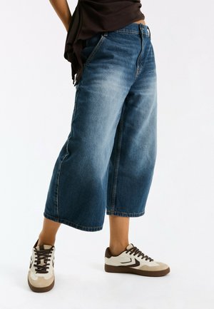 Person trägt weite, cropped blaue Jeans und beige Sneakers mit braunen Details auf weißem Hintergrund.