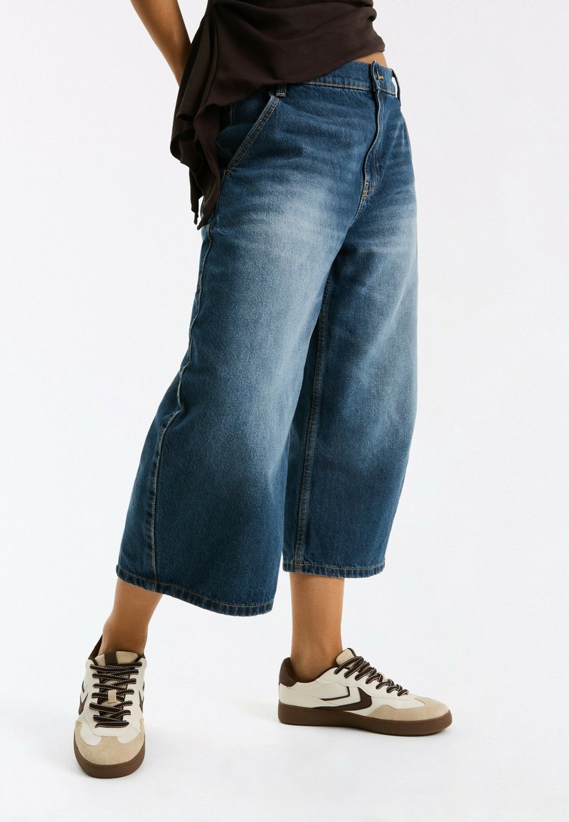 Personne portant un jean bleu large raccourci et des baskets beiges avec des détails marron sur fond blanc.