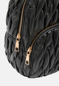 Primer plano de un bolso de cuero negro acolchado con dos cremalleras doradas y tiradores de cremallera grabados.