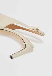 Beige høyhælte sko med slingback-reim og gullspenne. Glatt skinn, spiss tå og svart akcentert hæl.