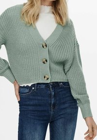 Cárdigan de punto verde claro con escote en V, que presenta tres botones grandes, puños acanalados y un diseño corto, combinado con jeans azul oscuro.