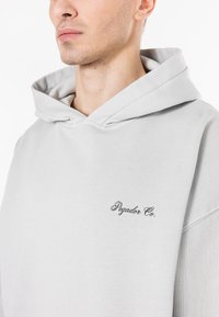 Hoodie cinza-claro com um tecido texturizado, apresentando um bolso frontal e um logotipo "Pegador Co." bordado em fio escuro.