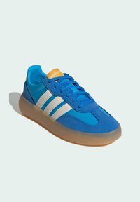 Plava atletska cipela s gornjim dijelom od brušene kože i sintetičkog materijala, s bijelim prugama, narančastim detaljima na jeziku i prozirnim đonom.
