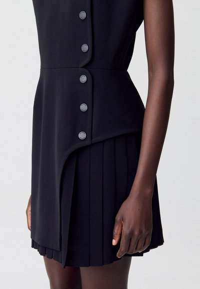 Un primo piano di un vestito nero senza maniche con un corpetto buttonato e una gonna plissée, mostrato accanto a una mano di pelle scura.