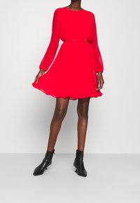 Punainen, poimutettu mini-mekko pitkillä hihoilla ja vyötäröltä kireällä leikkauksella, jossa on yhteensopiva vyö. Yhdistetty mustiin nilkkasaappaisiin.