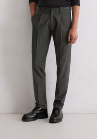 Grijze, op maat gemaakte broeken van gestructureerde stof, met plooien en een knoopsluiting, in combinatie met zwarte leren schoenen.