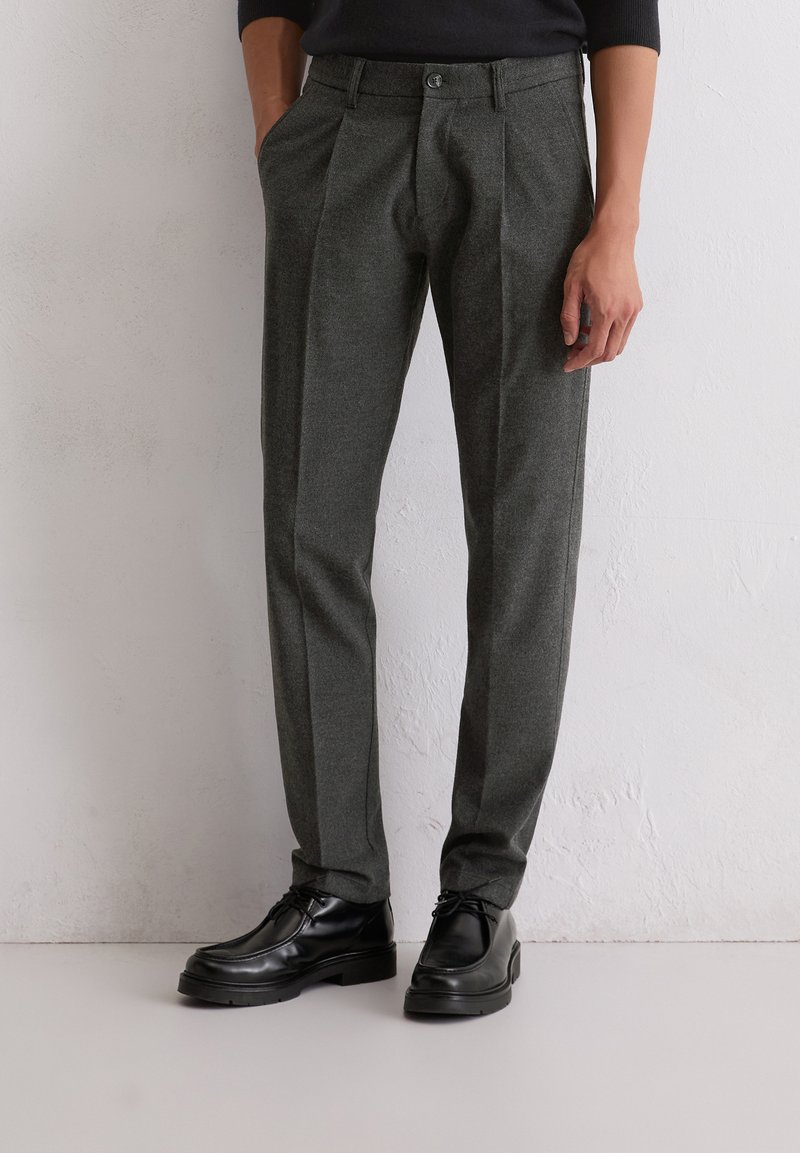 Grijze, op maat gemaakte broeken van gestructureerde stof, met plooien en een knoopsluiting, in combinatie met zwarte leren schoenen.