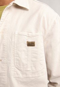 Beige canvas jacket featuring a front pocket with a branded patch. Includes a classic collar and clean seam detailing.
