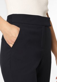 Pantaloni neri con una texture liscia, dotati di una vita ampia e tasche laterali, parzialmente coperti da una maglia chiara.
