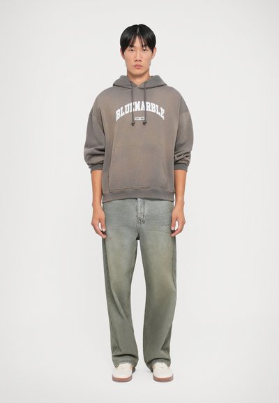 Sweat à capuche gris avec le texte "BLUIMARBLE", poche avant et cordons de serrage ; jeans baggy verdâtres ; baskets blanches avec des semelles brunes.