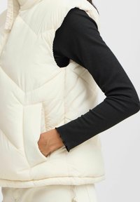 Dikke crème vest met hoge kraag, voorzien van zijzakken en chevronstiksels; gedragen over een zwart geribbeld langemouwen shirt.