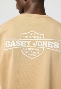 Camiseta de color tostado con cuello redondo que presenta texto impreso en blanco en la parte trasera, incluyendo "CASEY JONES" y "PARA TRABAJADORES," en un estilo retro y en negrita.