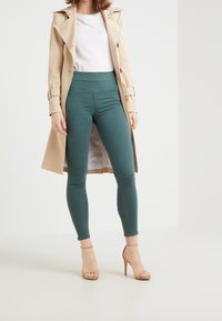 Beżowy płaszcz Trench na białej bluzce i tefowe, wysokie na talii wąskie spodnie, w połączeniu z cielistymi sandałami na wysokim obcasie. Gładki materiał i dopasowany krój.