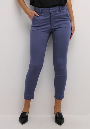 Vrouw draagt slanke, enkelhoge blauwe broek met zakken, gecombineerd met zwarte loafers met puntige neus, staand tegen een witte achtergrond.