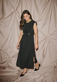 Čierne bezrukávové šaty s pásom v páse a okrúhlou kovovou aplikáciou, bočný rozparok a hladká tkanina. Nosené s čiernymi špicatými topánkami.