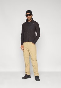 Chaqueta polar negra con cremallera, tejido texturizado y bolsillo en el pecho, combinada con pantalones color caqui y zapatos negros, llevada con un gorro negro y gafas de sol.