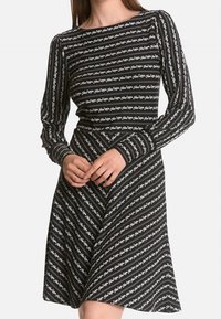 Schwarzes Langarmkleid mit einem weißen Blumenstreifenmuster, tailliertem Oberteil und ausgestelltem Rock. Das Material wirkt glatt und dehnbar.