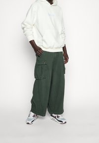 Weißer Fleece-Hoodie mit Fronttasche, kombiniert mit dunkelgrünen, weit geschnittenen Cargo-Hosen mit Seitentaschen. Schuhe: grau-rote Sneaker.