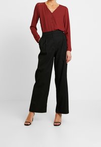 Blouse boutonnée bordeaux avec détails en dentelle et manches longues, associée à un pantalon noir taille haute. Sandales à brides noires complètent la tenue.