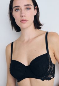 Sutiã de renda preta com uma forma estruturada, suporte de aro e alças ajustáveis. Apresenta tecido texturizado e bordas de renda em formato de concha.